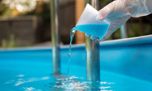 Hand holding and pouring algicide blue liquid from glass into water of swimming pool. Water purification and prevention of appearance of algae and microorganisms.